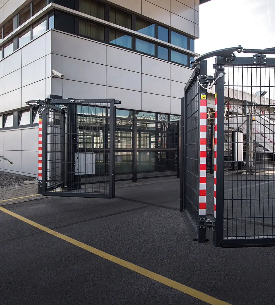 Antracytowa brama składana rozłożona na wjeździe na teren zakładu — skrzydła z siatki na prowadnicy górnej, słupki w biało-czerwone pasy